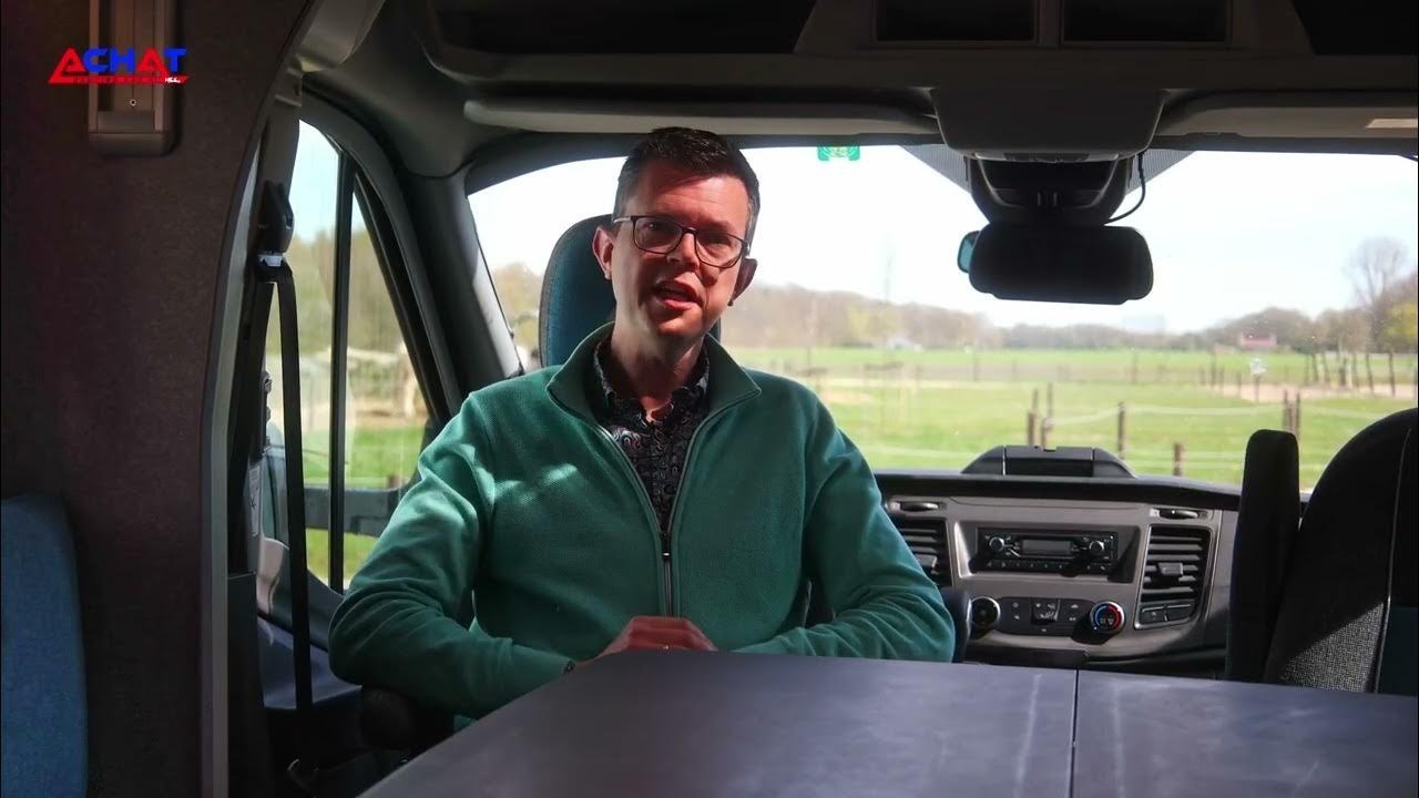 Un homme portant des lunettes et une veste turquoise est assis à l'intérieur d'un camping-car, peut-être considéré comme un achat camping car, avec des champs visibles à travers le pare-brise avant. La lumière pénètre à l'intérieur, éclairant une partie du poste de conduite et du tableau de bord.