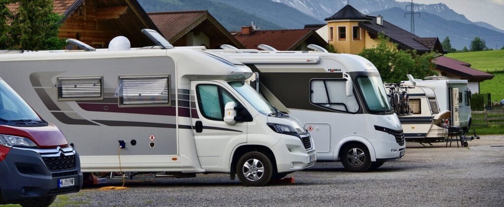 Plusieurs camping-cars et fourgonnettes de camping sont garés côte à côte dans un terrain près de maisons et de champs verts, avec des montagnes visibles en arrière-plan sous un ciel partiellement nuageux.