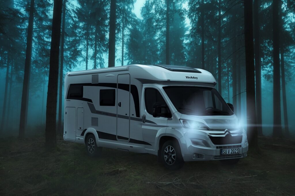Un camping-car blanc avec les phares allumés est garé dans une forêt dense et brumeuse entourée de grands arbres sous un ciel crépusculaire.