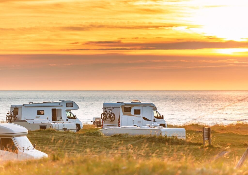 Trois camping-cars sont garés sur un terrain herbeux au bord de la mer au coucher du soleil, avec un ciel doré se reflétant sur l'eau. L'un d'eux est équipé de vélos à l'arrière. Le décor évoque une atmosphère paisible de camping côtier.