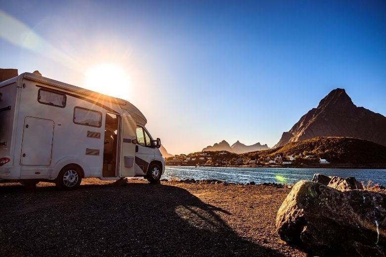 Un camping-car garé au bord d'un plan d'eau calme au coucher du soleil, avec des montagnes en arrière-plan et le soleil qui brille. Des rochers sont visibles au premier plan.