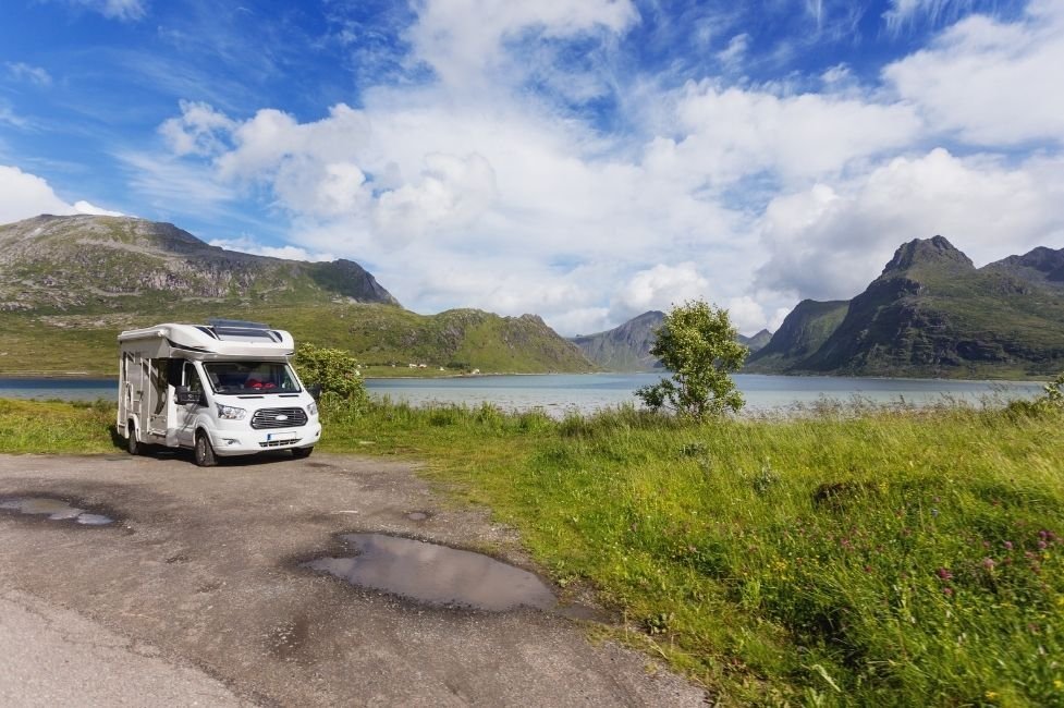 Un camping-car blanc est garé au bord d'un lac, entouré d'herbe verte et de montagnes, sous un ciel partiellement nuageux. Le paysage est paisible et pittoresque, évoquant un lieu isolé et naturel, idéal pour le camping.