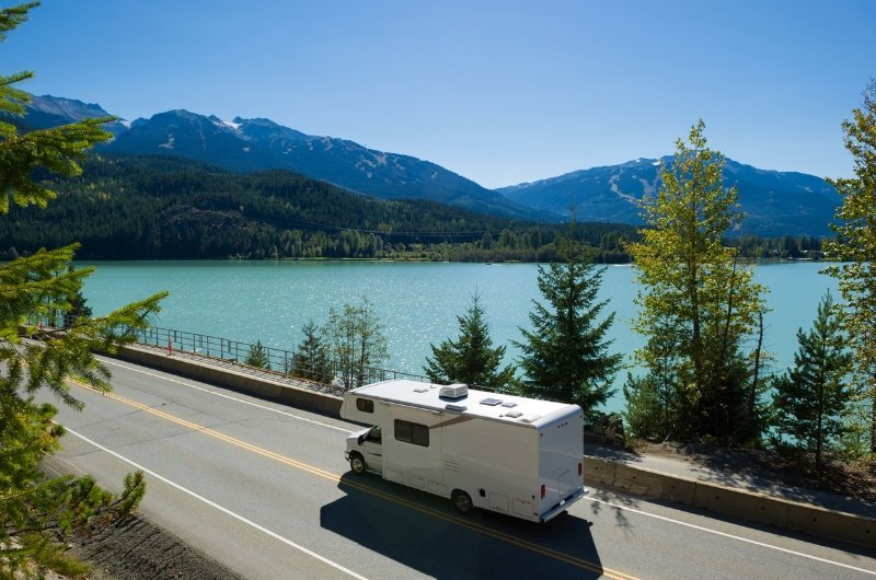 Un camping-car blanc roule le long d'une route au bord d'un lac bordée de pins, avec de l'eau turquoise et des montagnes boisées en arrière-plan sous un ciel bleu clair.