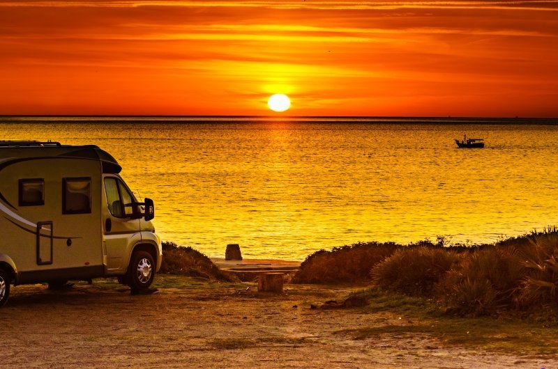 Un camping-car est garé au bord d'une plage, surplombant une mer calme au coucher du soleil. Le ciel s'illumine d'orange et de jaune tandis que le soleil se couche à l'horizon, laissant entrevoir un petit bateau sur l'eau.