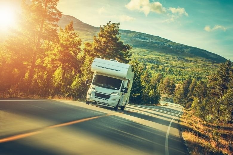 Un camping-car blanc roule sur une route pittoresque et sinueuse entourée d'arbres et de montagnes sous un ciel lumineux et ensoleillé.