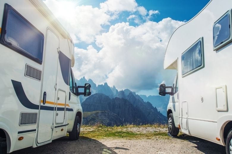 Deux camping-cars blancs garés sur une route de montagne avec une vue panoramique sur des sommets déchiquetés au loin sous un ciel partiellement nuageux et un soleil éclatant.
