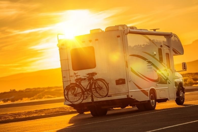 Un camping-car blanc avec des vélos montés à l'arrière est garé sur une route vide au coucher du soleil, avec la lumière dorée du soleil qui coule en arrière-plan et projette de longues ombres.