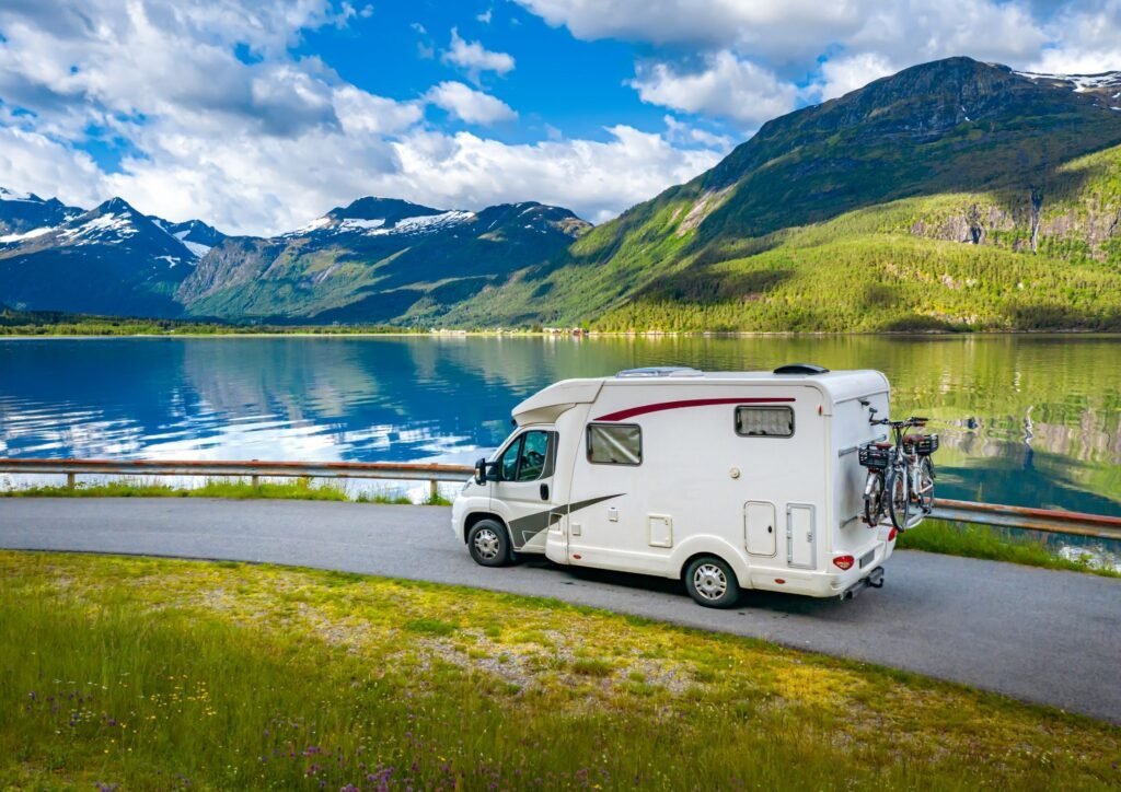Un camping-car blanc est garé au bord d'un lac pittoresque, entouré d'herbe verte, de montagnes et d'un ciel bleu parsemé de nuages. Deux vélos sont installés à l'arrière.